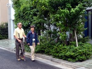白杖を使っている男性が女性ガイドヘルパーと道路を歩いている写真
